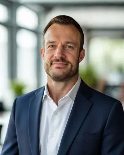 Smiling man in a navy blue suit and white shirt in a bright modern office setting with blurred background.