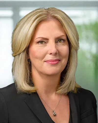 Professional woman with blonde hair wearing black blazer and silver necklace in a bright office setting
