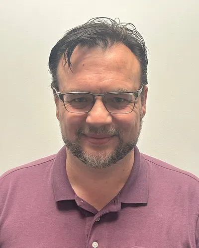 Middle-aged man with glasses and beard smiling, wearing a purple polo shirt against a plain background