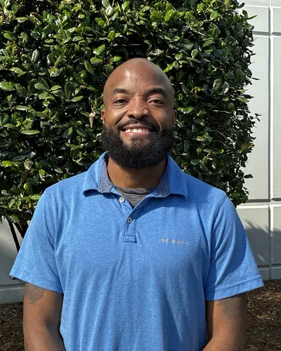 Smiling bald man with a beard wearing a blue Columbia polo shirt standing outdoors near greenery and a building.