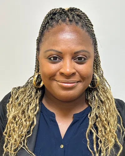 Smiling woman with braided blonde hair wearing gold hoop earrings and a navy blue top against a plain background