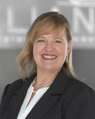 Smiling woman wearing a pinstripe suit jacket, white blouse, pearl necklace, and gold earrings in a professional setting.