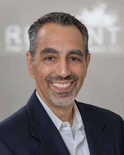 Smiling middle-aged man with salt and pepper hair wearing a dark blazer and white shirt against a gray background.