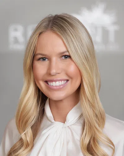 Smiling blonde woman with long hair wearing a white blouse with a tied bow on a neutral background.