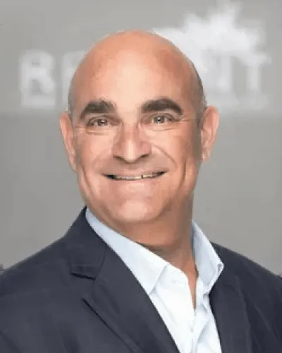 Smiling bald man in a dark suit and white shirt posing against a gray background with blurred text.