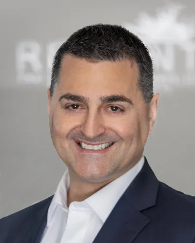 Portrait of a smiling man in a suit jacket and white shirt against a blurred gray background.