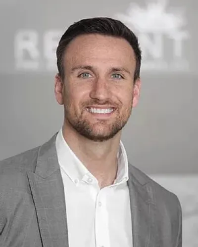 Smiling man with blue eyes wearing a gray suit jacket and white shirt against a blurred background.