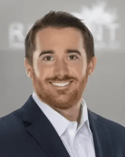 Smiling man with brown hair and beard wearing a dark suit jacket and white shirt against a gray background.