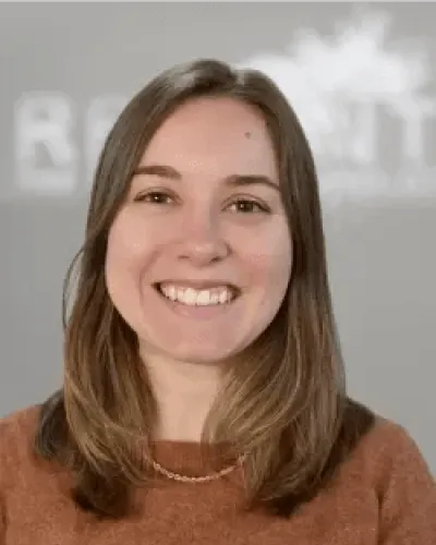 Young woman with shoulder-length light brown hair smiling while wearing a brown sweater and necklace indoors
