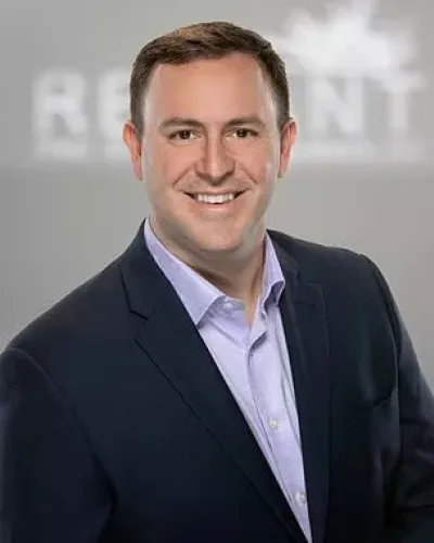 Smiling man in dark blazer and light shirt against blurred gray background with partial text.