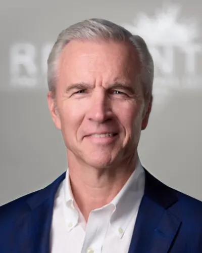 Smiling mature man with gray hair wearing a white shirt and blue blazer against a blurred logo background.