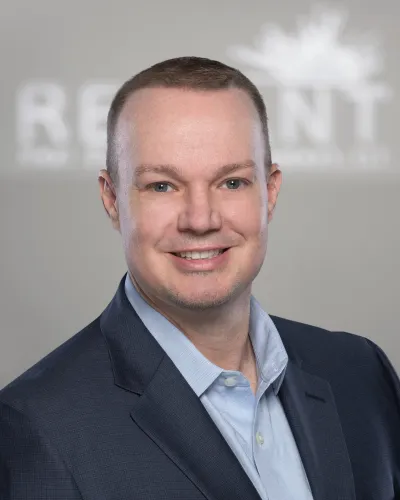 Smiling man in a blue dress shirt and dark blazer with a blurred office logo background