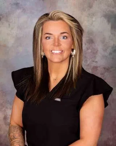 Smiling woman with blonde and brown hair wearing black top with ruffled sleeves and earrings against mottled gray background