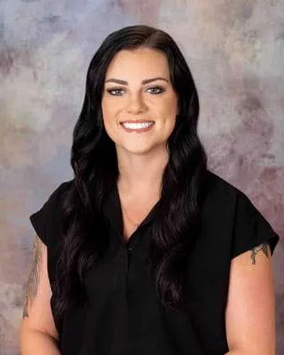 Smiling woman with long black hair and tattoos wearing a black shirt against a neutral background