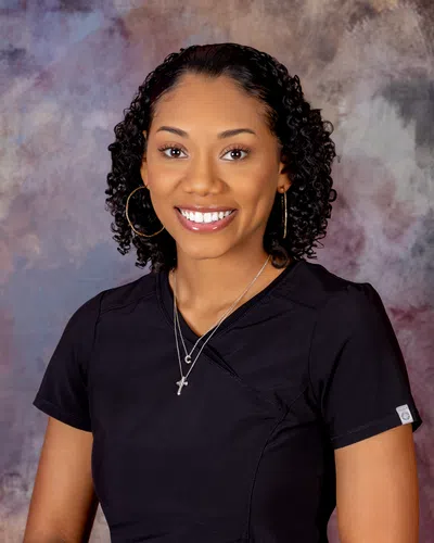 Smiling woman with curly hair wearing black scrubs and hoop earrings against a soft textured background