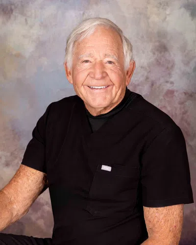Smiling elderly man with white hair wearing black medical scrubs sitting against a neutral textured background