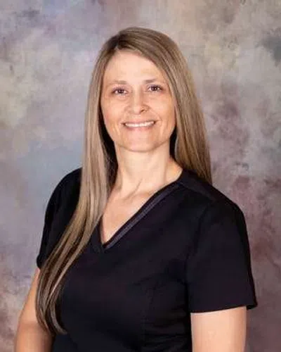 Smiling woman with long straight hair wearing black scrub top against a neutral studio background