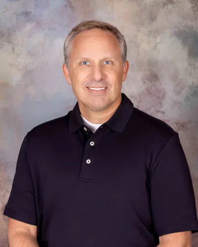 Smiling middle-aged man wearing a black polo shirt against a soft, mottled gray and beige background.