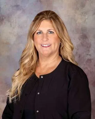 Smiling woman with long blonde hair wearing a black top against a blurred neutral background.