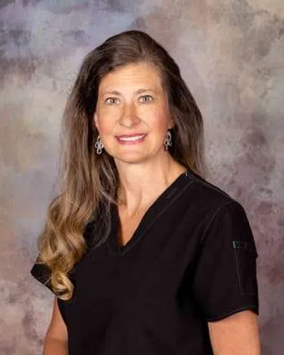 Woman with long wavy hair wearing black medical scrubs and silver earrings smiling in front of a neutral backdrop