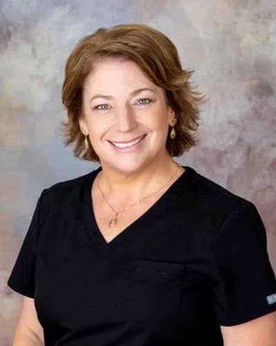 Smiling woman with short brown hair wearing black scrubs and gold jewelry against a neutral background