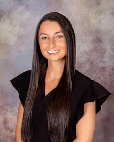 Smiling young woman with long straight brown hair wearing a black top against a textured gray background