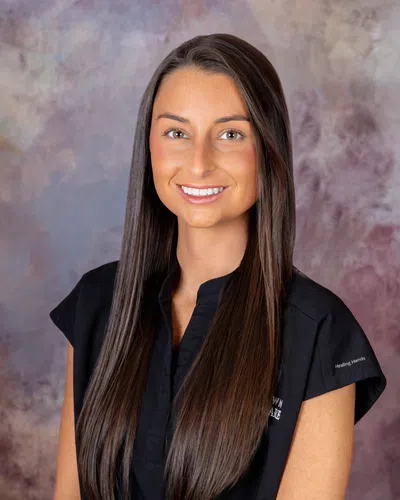 Young woman with long brown hair wearing a black shirt smiles against a soft purple and gray background.