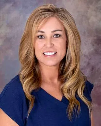 Smiling woman with long blonde hair wearing a blue top against a blurred gray and purple background