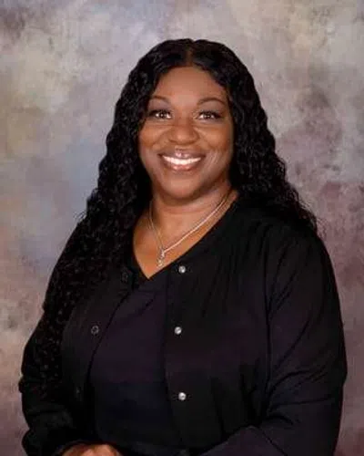 Smiling woman with long curly hair wearing a black top and jacket against a textured gray background