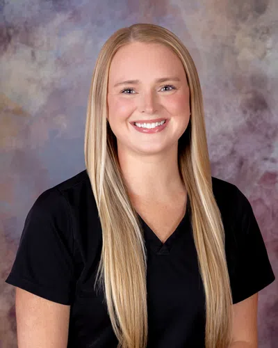 Smiling woman with long blonde hair wearing a black scrub top against a mottled background