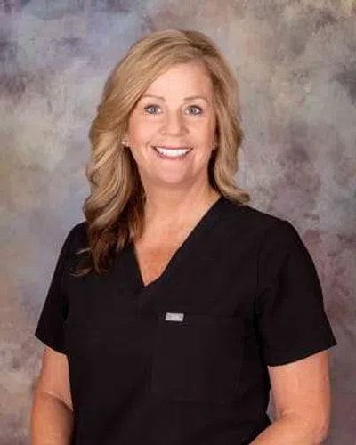 Smiling woman with long blonde hair wearing black medical scrubs against a textured background