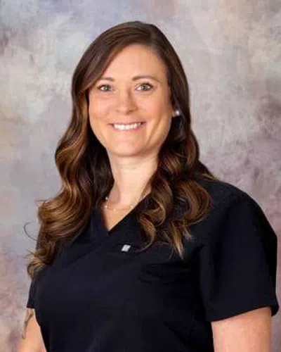 Smiling woman with long brown hair wearing black medical scrubs against a neutral backdrop