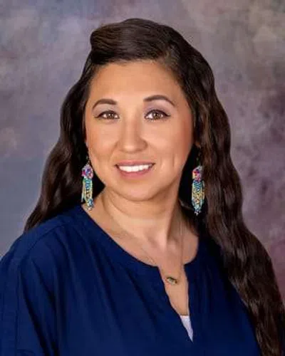 Smiling woman with long dark hair, wearing blue top and colorful dangling earrings against a blurred background.