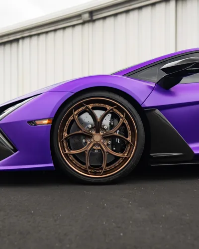Close-up of a purple sports car front wheel with intricate bronze alloy rim and black tire on asphalt.