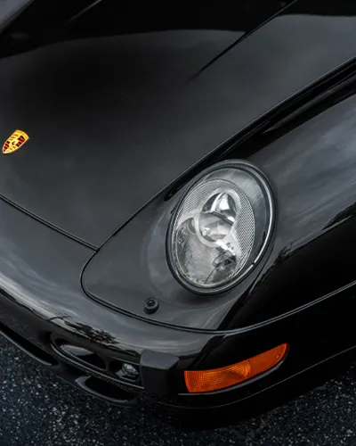Close-up of a black Porsche sports car front headlight and emblem on hood under natural light.