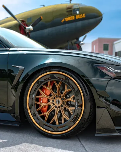 Close-up of a sleek black sports car with custom bronze and gold rims parked near a vintage plane on a sunny day.