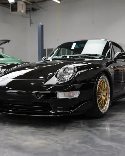 Shiny black sports car with gold rims parked inside a modern garage with another green car nearby.