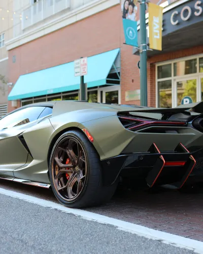 Matte green luxury sports car with bronze wheels parked on urban street near brick building and blue awning