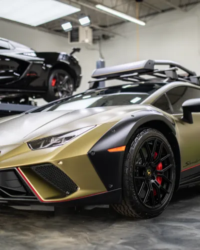 Matte gold Lamborghini Huracan with black widebody kit and roof rack in a modern garage setting.