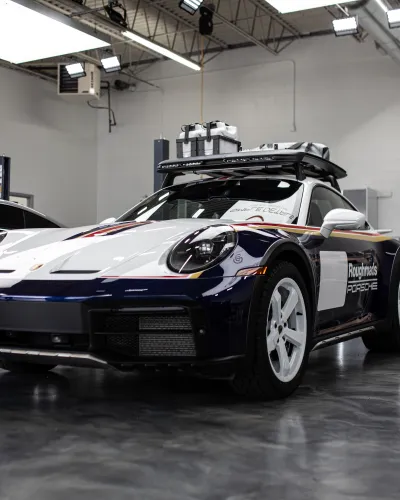 White and navy blue Porsche sports car with roof cargo box in an indoor garage with polished floor.