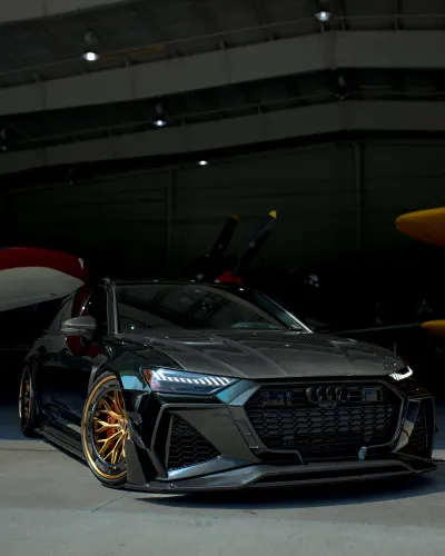 Sleek black Audi car with golden rims parked inside an aircraft hangar near vintage planes.