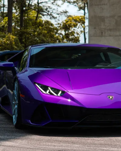 Purple Lamborghini sports car parked outdoors with a black sedan and trees in the background under sunlight.