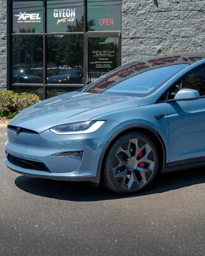 Blue Tesla Model X parked outside a car detailing shop with signage and an open door.