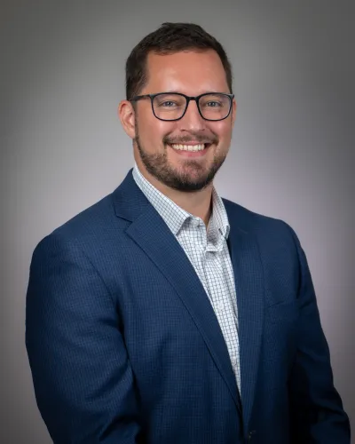 Smiling man with glasses wearing a blue suit jacket and white checkered shirt against a neutral background