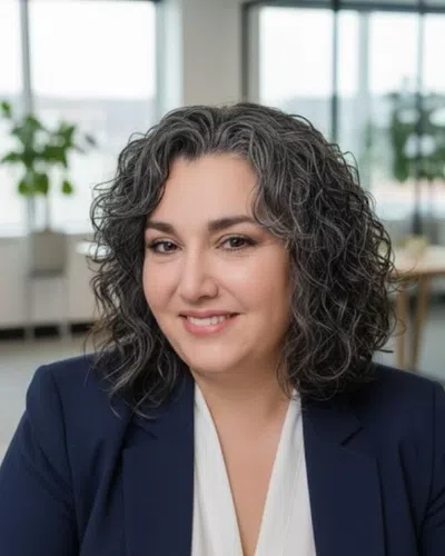Smiling professional woman with curly salt-and-pepper hair wearing a navy blazer in a modern office.