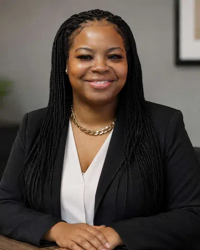 Confident woman with braided hair in black blazer smiling, sitting at a desk in a modern office environment.