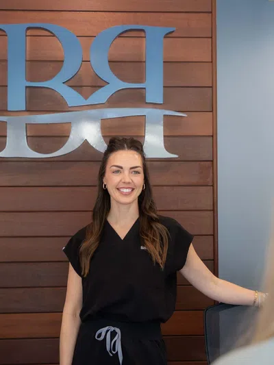 Smiling woman in black uniform standing in front of wooden wall with silver RRH logo in office setting
