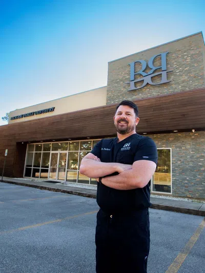 Smiling man in black scrubs stands with arms crossed outside modern dental office building under clear blue sky
