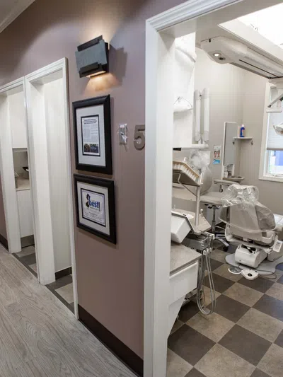 Bright dental clinic hallway with open patient rooms and modern dental equipment in one room visible on checkered floor.