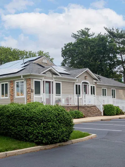 Single-story house with solar panels on the roof surrounded by greenery and an empty parking lot under a blue sky.
